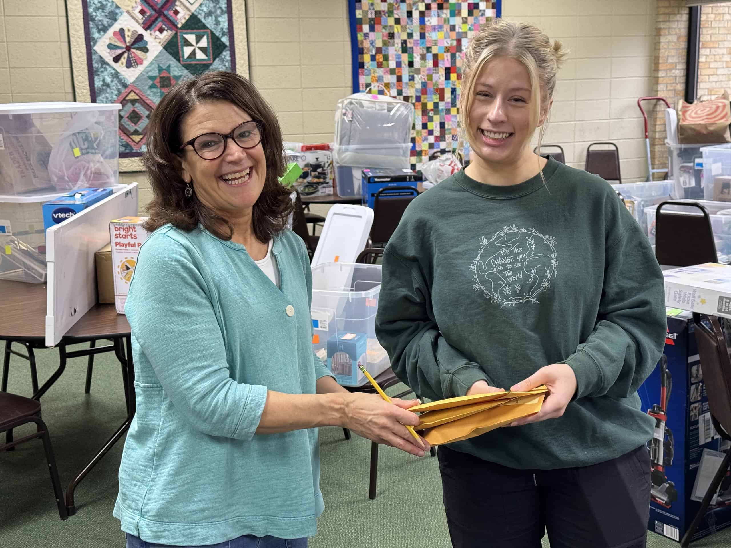 Two people smiling in a classroom setting with quilts and supplies around them. They hold envelopes together, engaging in a friendly interaction.
