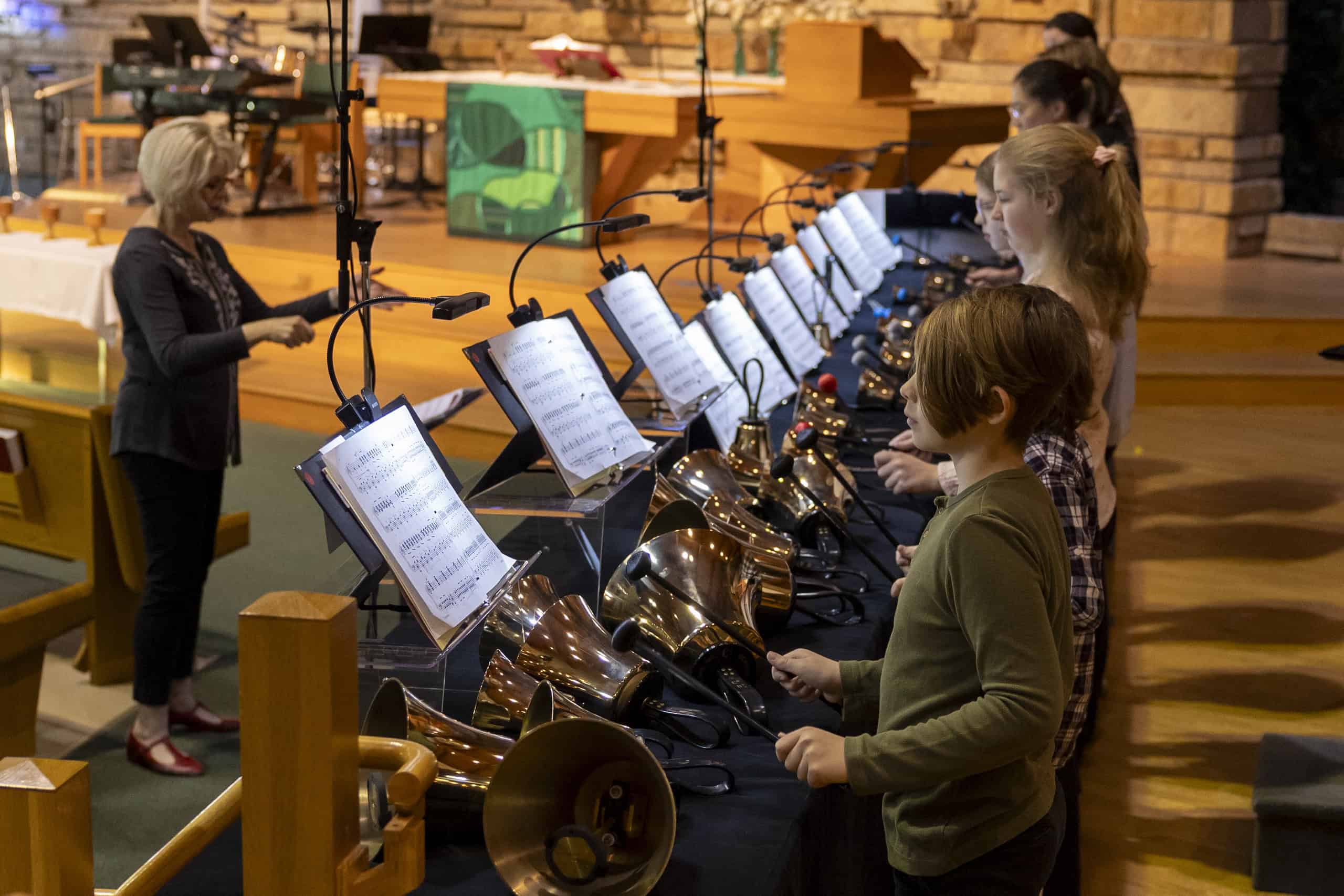 A person conducts children playing handbells in a church setting. Music stands with sheet music are in front of each child.