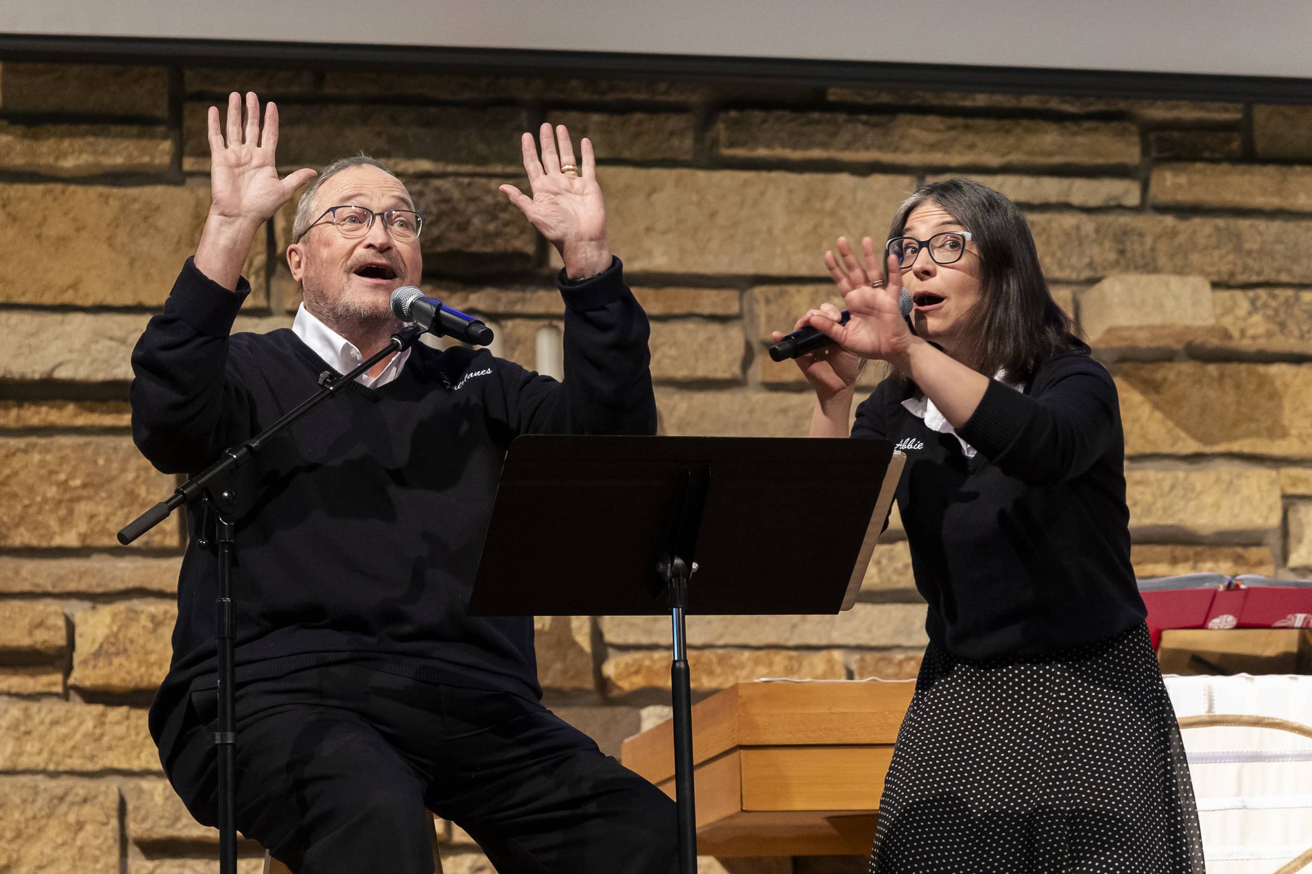 Two people enthusiastically speak into microphones on a stage against a stone wall backdrop, engaged in a lively presentation or performance.