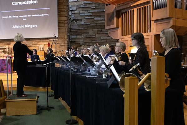 A group of people are playing handbells in a church setting, with a conductor and visible organ pipes in the background.