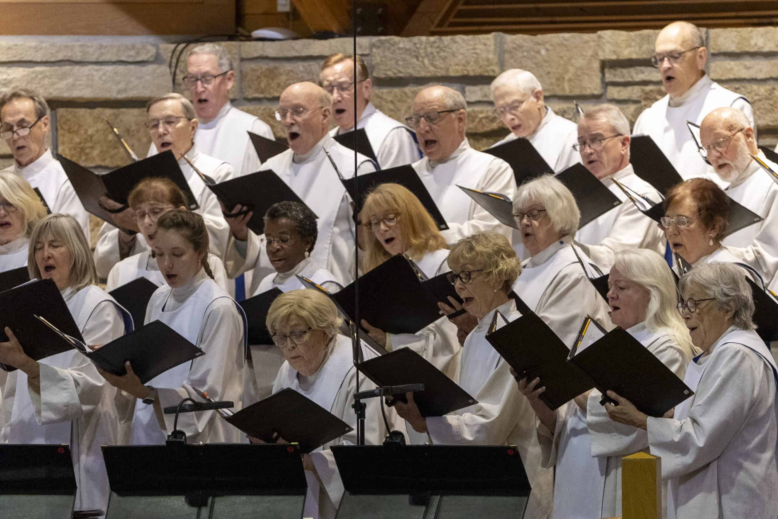 A choir of people in white robes sings passionately, holding black folders, standing against a stone wall in a church setting.