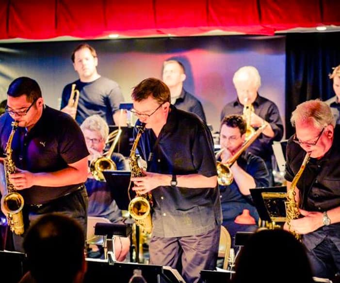 A group of people are playing saxophones and trumpets on stage, all wearing black shirts, under a red curtain in a small venue.