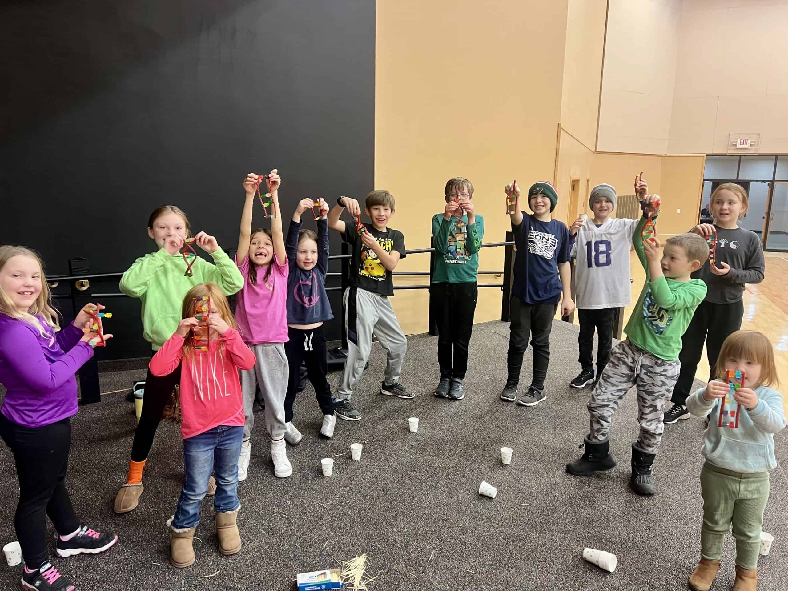Children indoors holding colorful crafts, standing on a carpeted floor. They appear happy and engaged, with scattered cups on the ground.