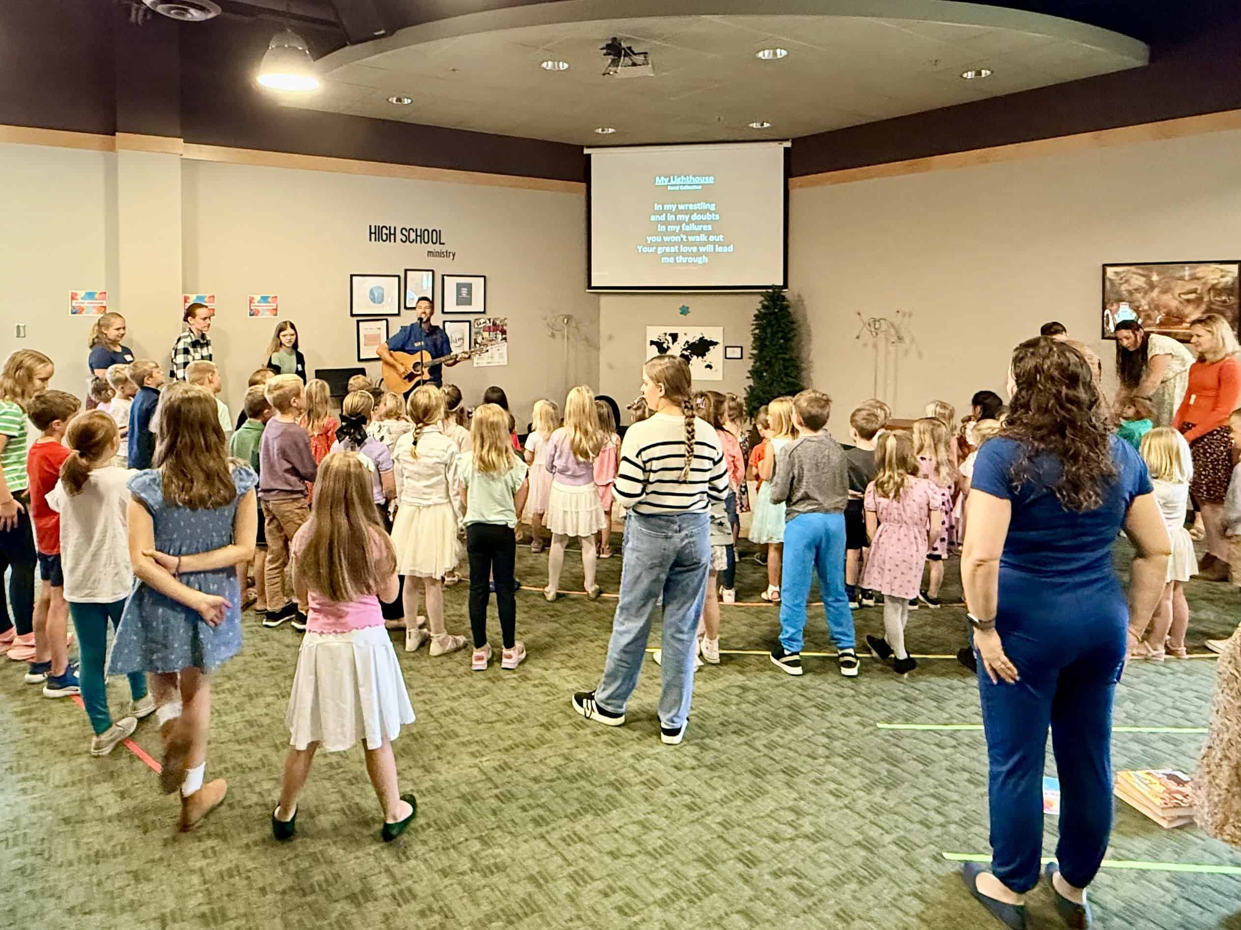 A group of children and a person with a guitar stand in a room labeled "High School Ministry" during a singing activity.