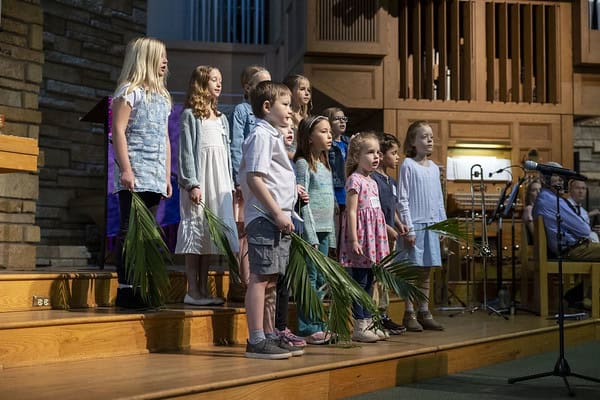 Children stand on a wooden stage holding palm fronds, singing in a church setting. A person is seated with a musical instrument.
