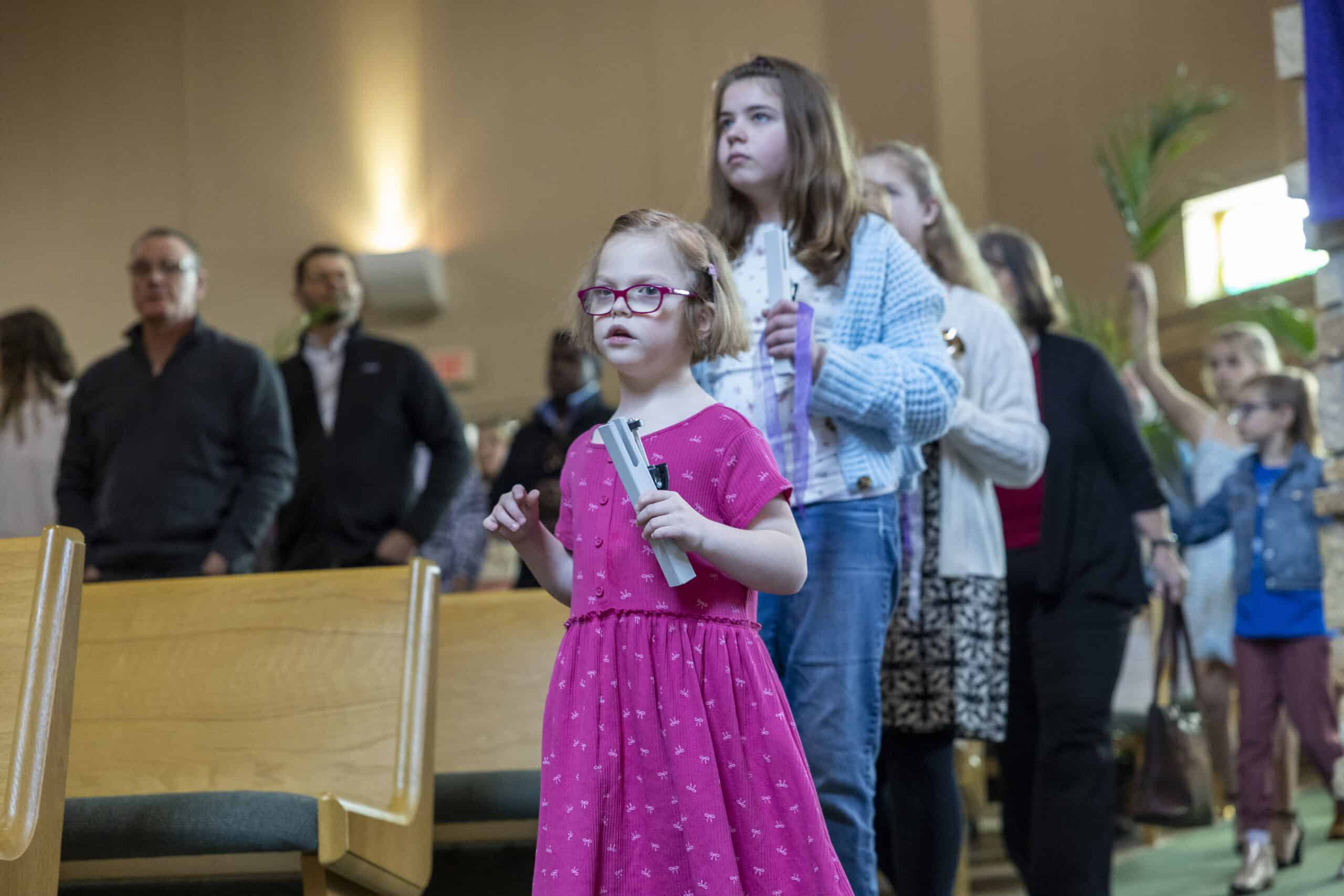 A group of people, including children holding items, stand inside a building with wooden pews and soft lighting.