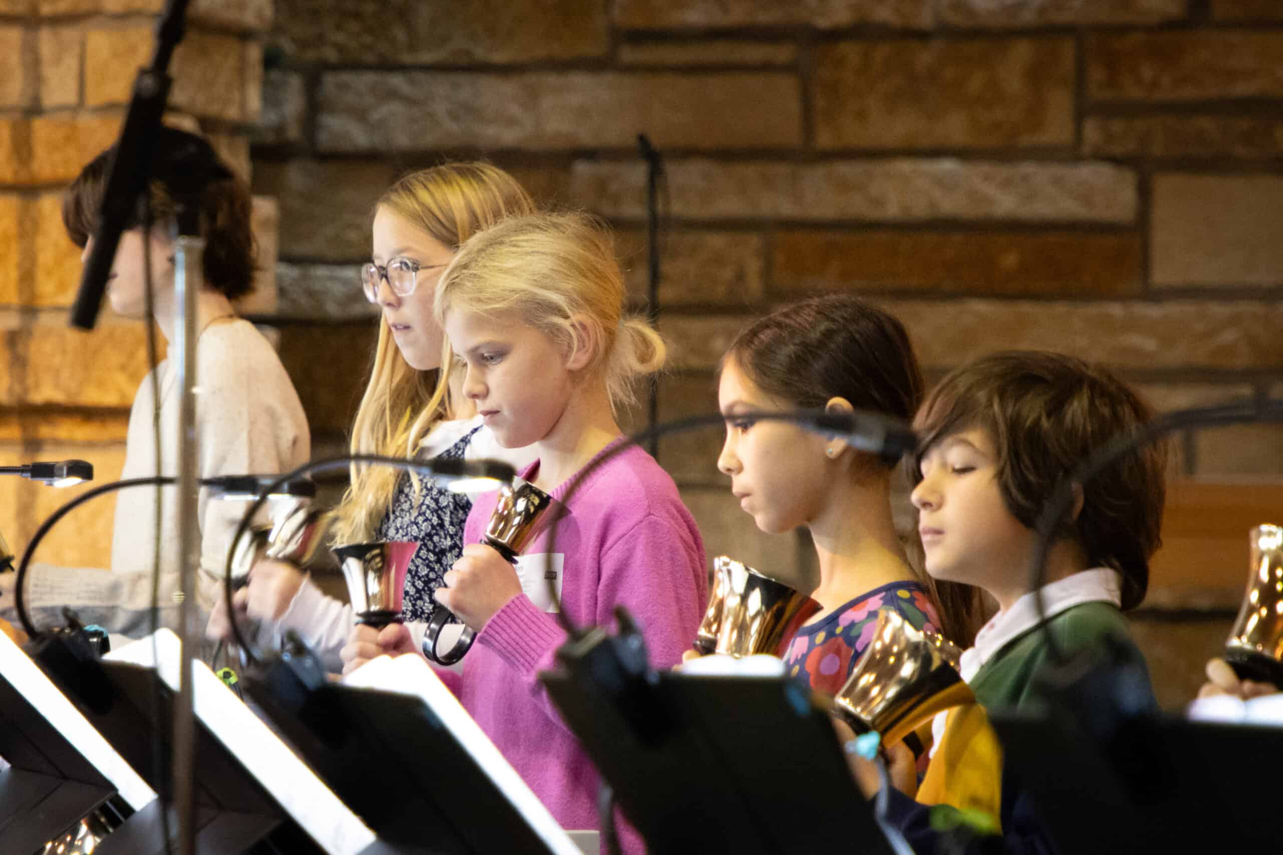 A group of children playing handbells in a church setting, with music stands and a stone wall in the background.