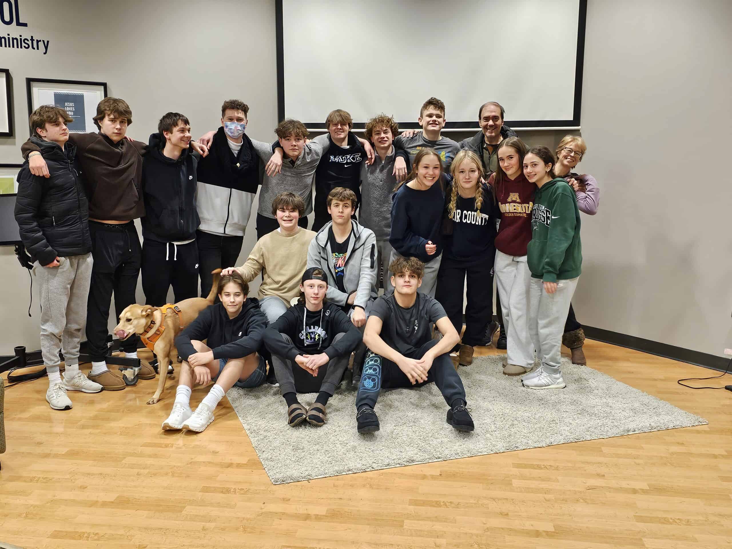 A group of young people and two adults pose indoors with a dog. Casual clothing and a warm setting suggest a friendly gathering.