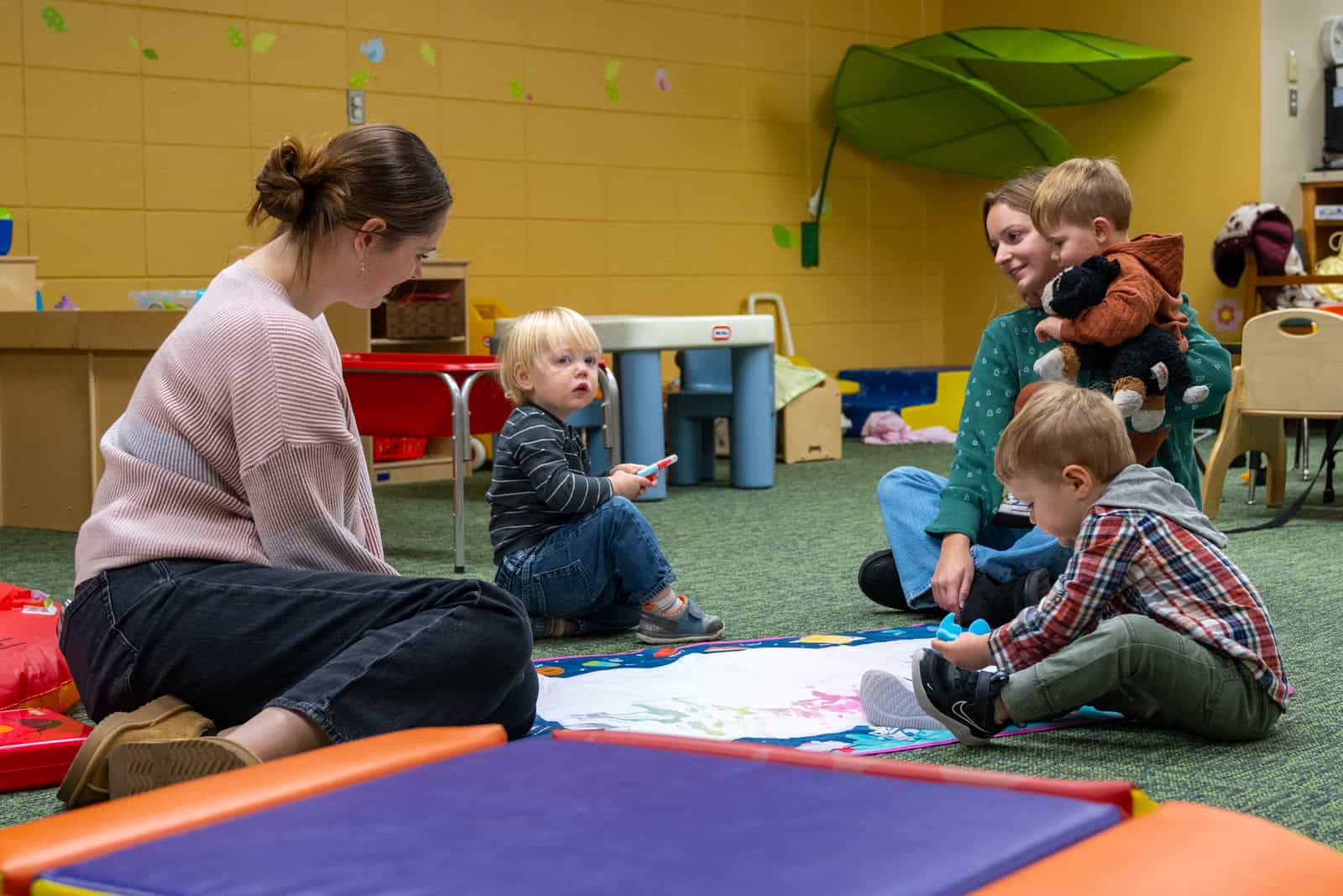 Two persons and three children are sitting on the floor of a brightly decorated playroom, engaging in activities together.