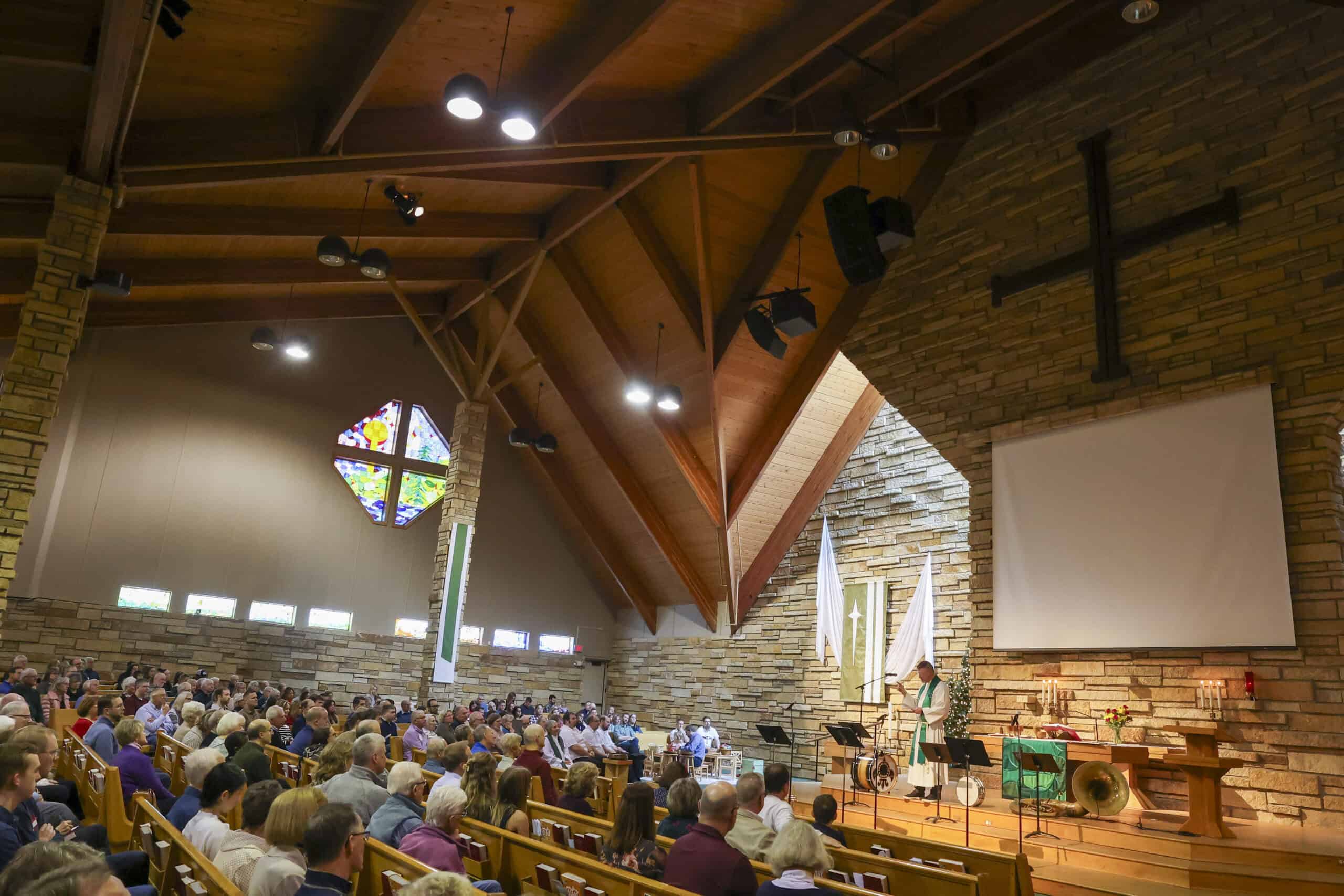 A church congregation gathered inside Incarnation Lutheran Church during a service or event, with a prominent cross on the brick wall and a large screen at the front.