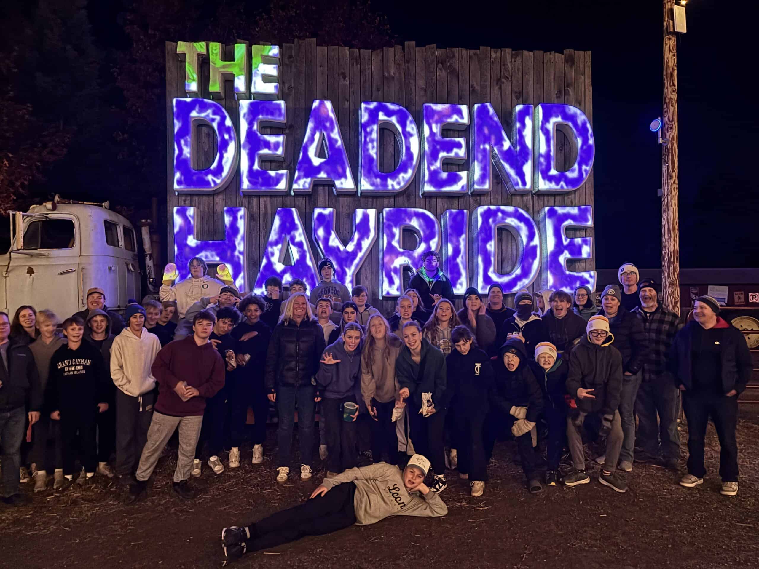 A detailed group photo of people gathered at night in front of a large illuminated sign reading "The Deadend Hayride," promoting a local haunted attraction.