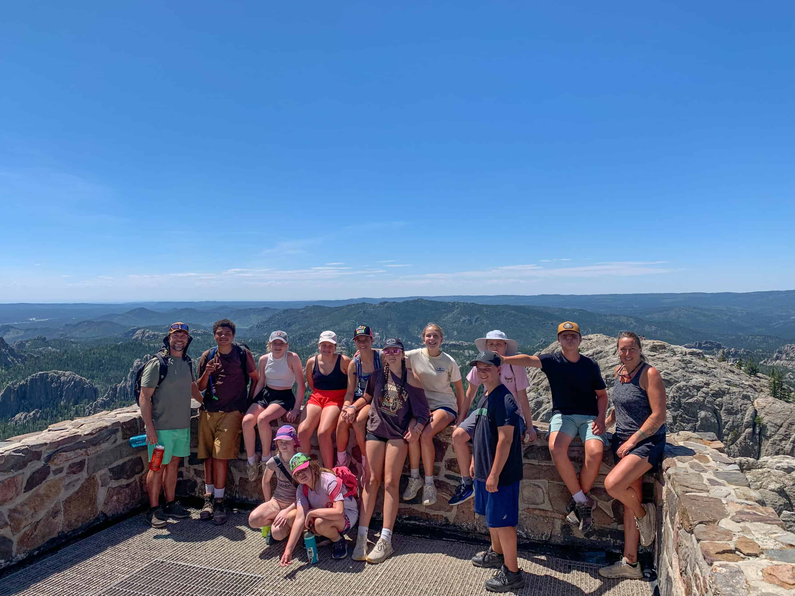 A detailed group of people, including children and adults, standing and sitting on a stone overlook with a scenic canyon and blue sky in the background, during daytime.