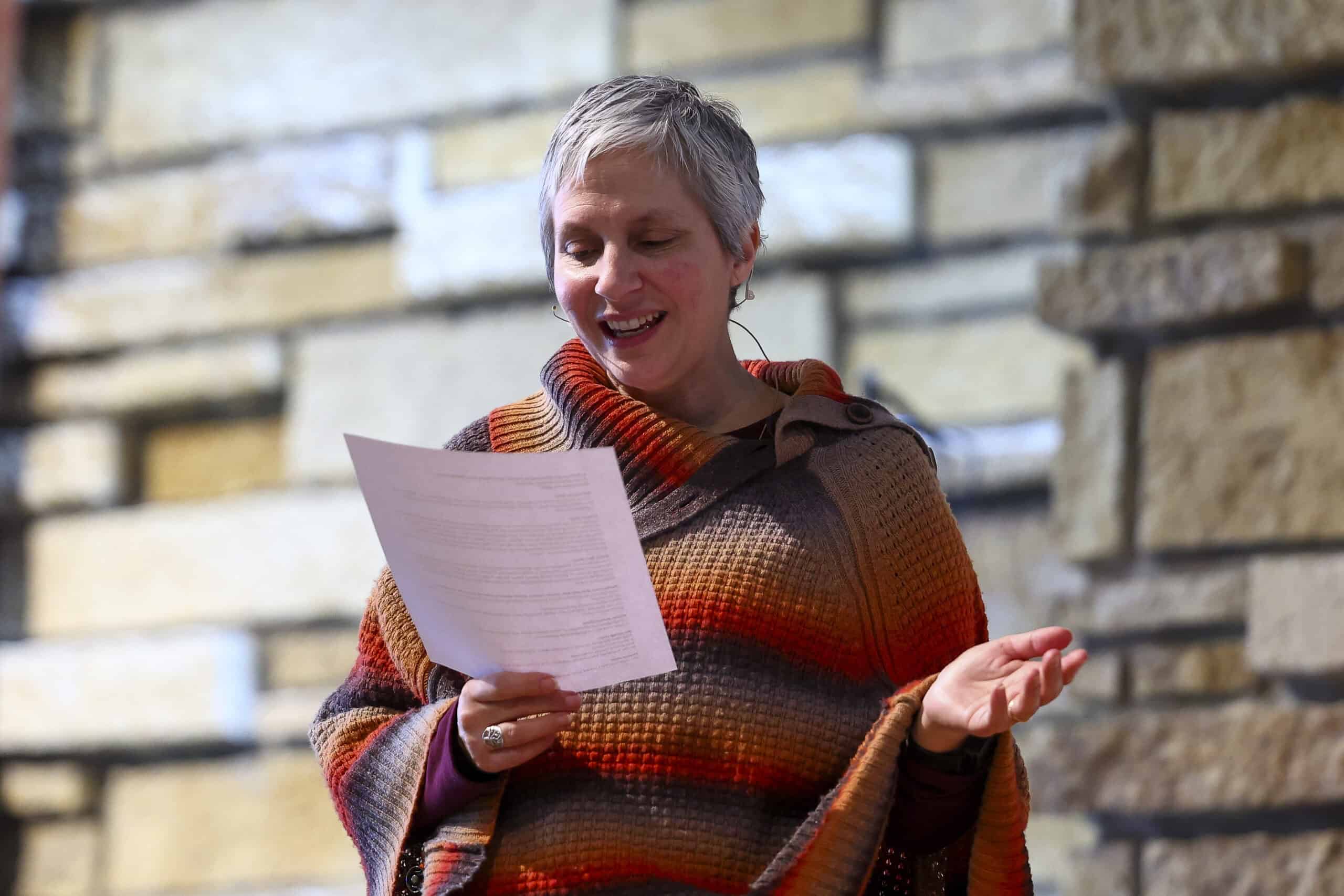 A woman smiling and reading from a paper in front of a stone wall at Incarnation Lutheran Church, engaging in a community or worship activity.