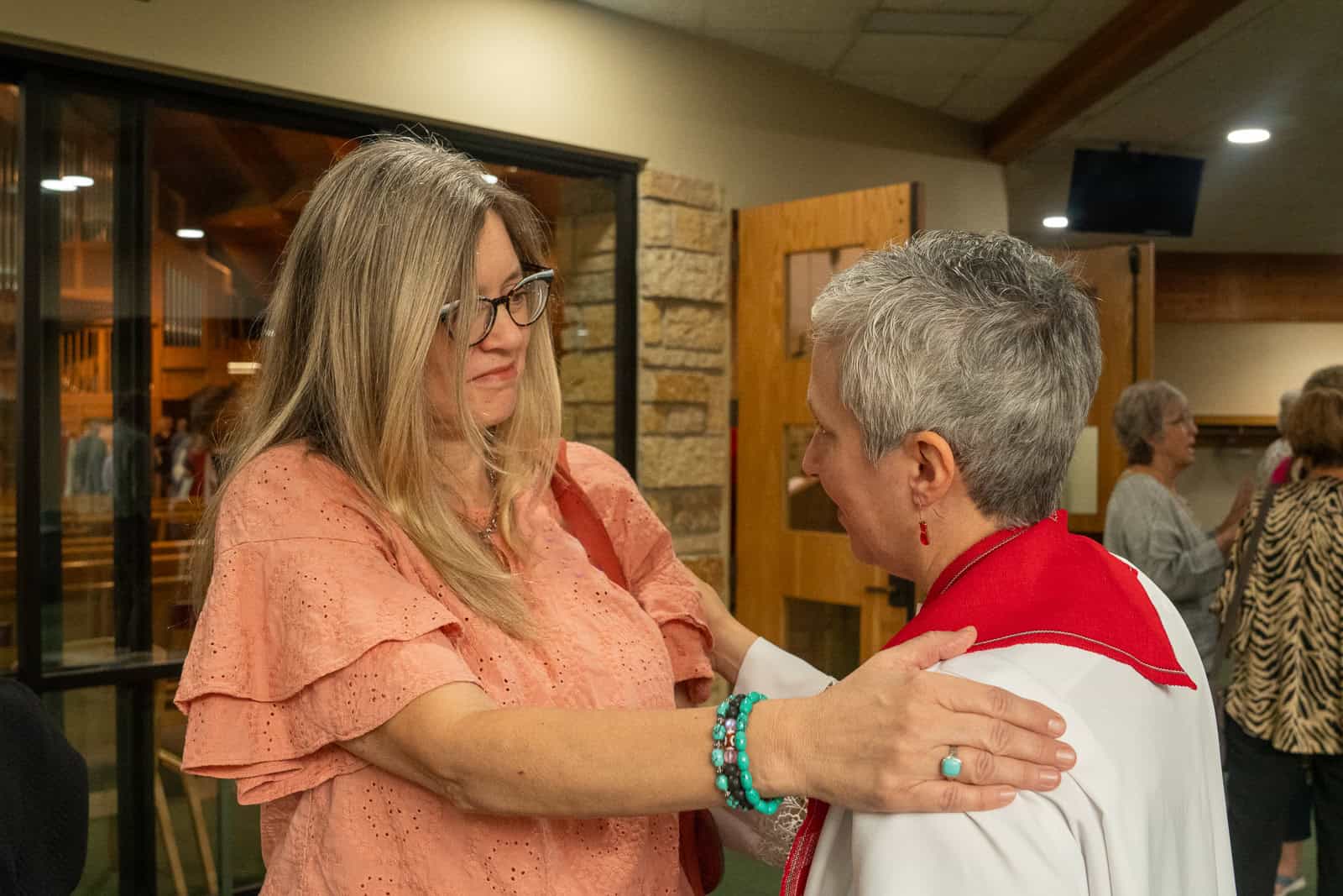 Care & Support Two women sharing a heartfelt moment at Incarnation Lutheran Church, one with glasses and long blonde hair, the other with short gray hair in a red and white outfit.