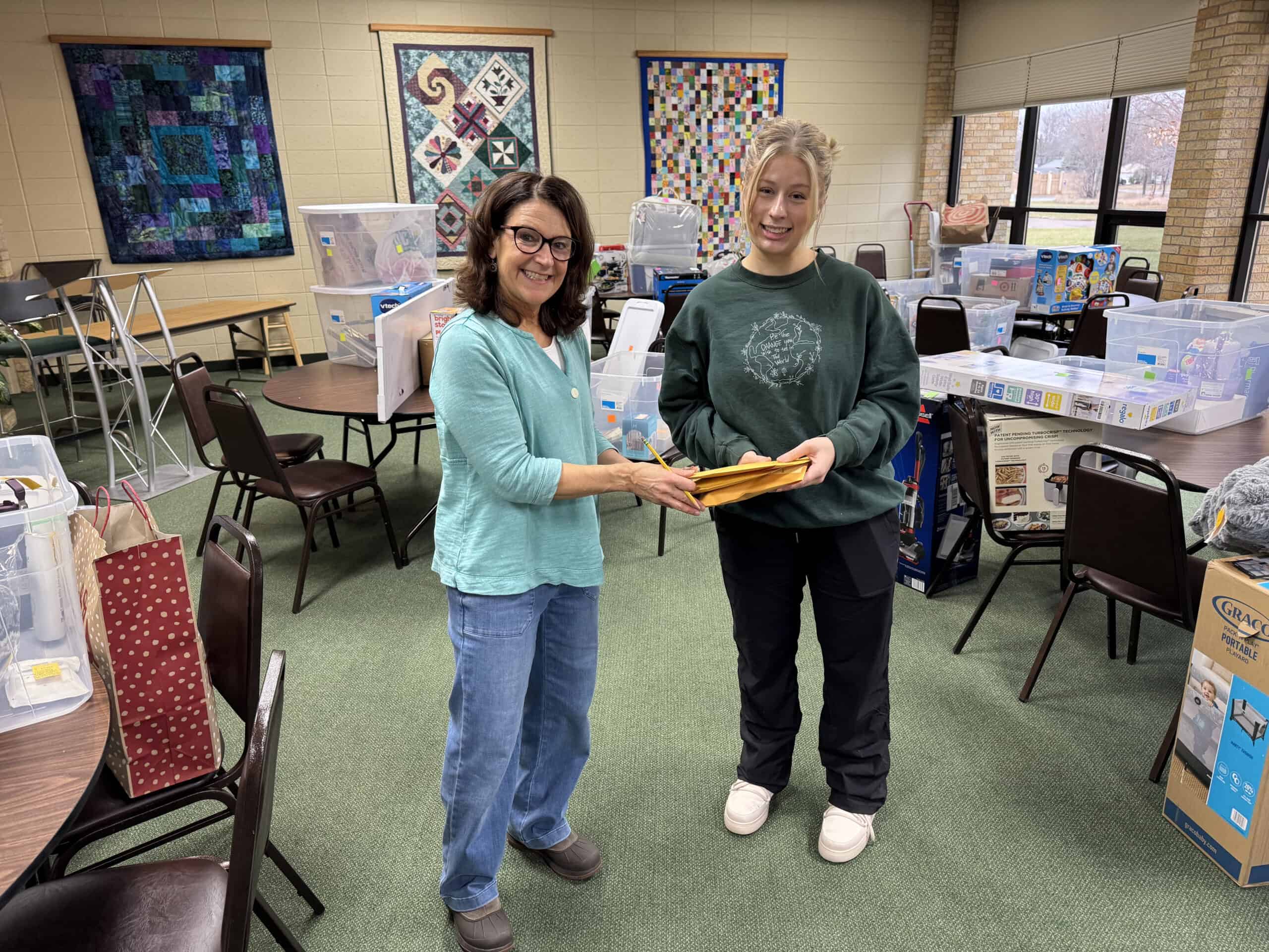 Two women smiling and exchanging a quilt in a quilting room at Incarnation Lutheran Church, with colorful quilts hanging on the wall behind them.