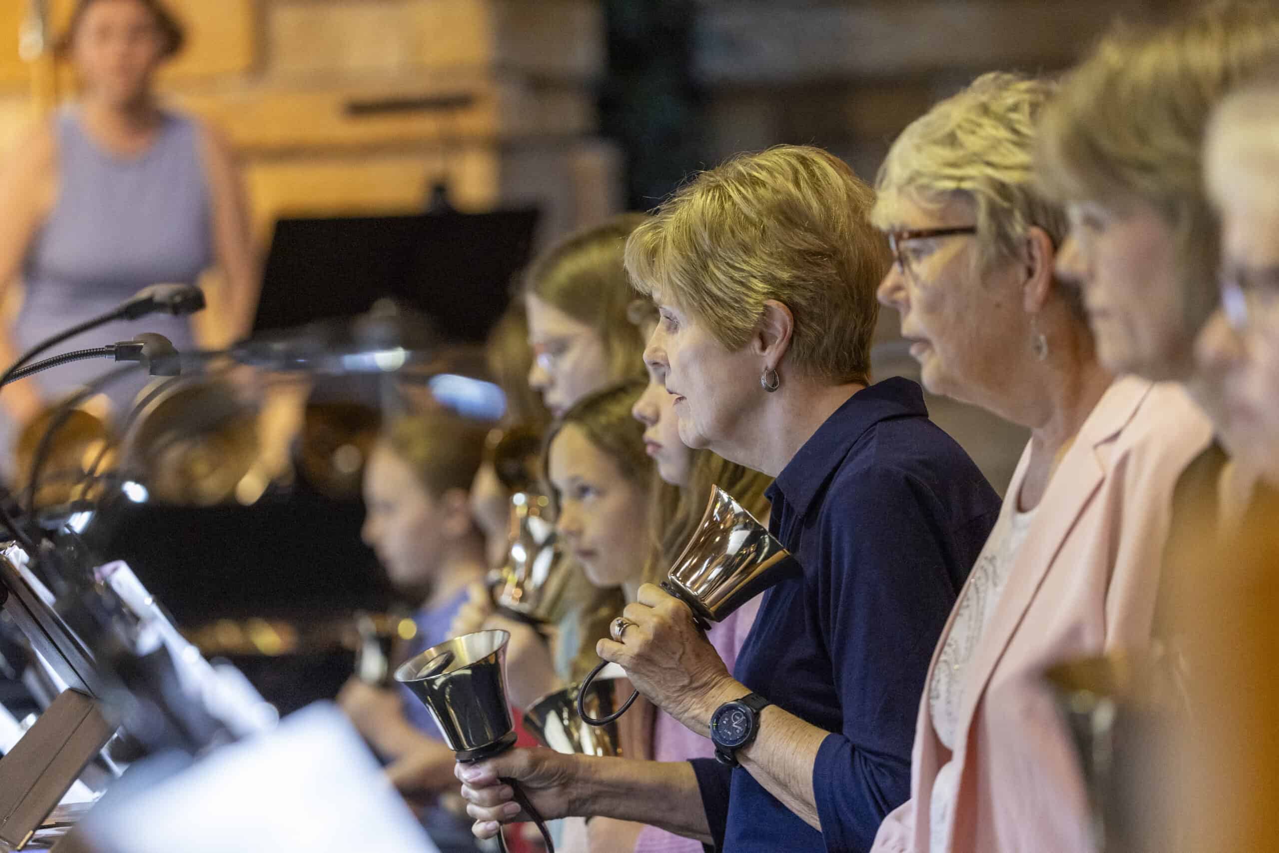 People playing handbells in a group setting, focusing on hands and instruments. The image captures the shiny, metallic handbells mid-performance.