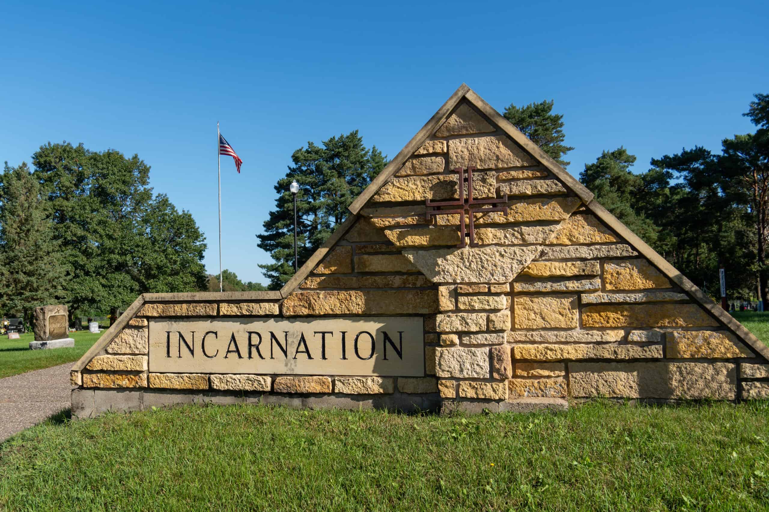 Stone sign for Incarnation Lutheran Church with a cross and American flag in the background, surrounded by trees and a clear blue sky.