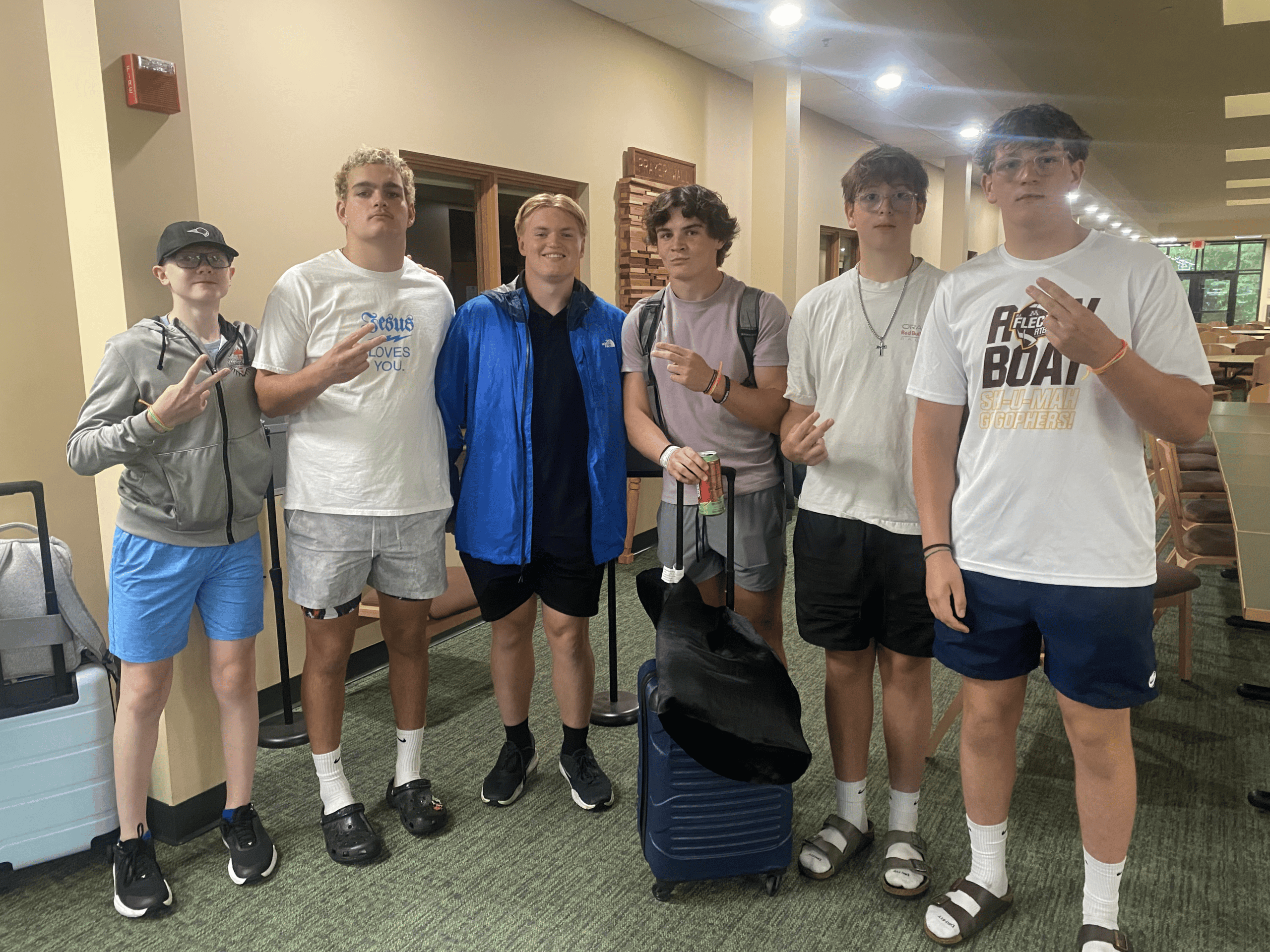 Group of six young people standing indoors, some with luggage, smiling and making peace signs, at Incarnation Lutheran Church.