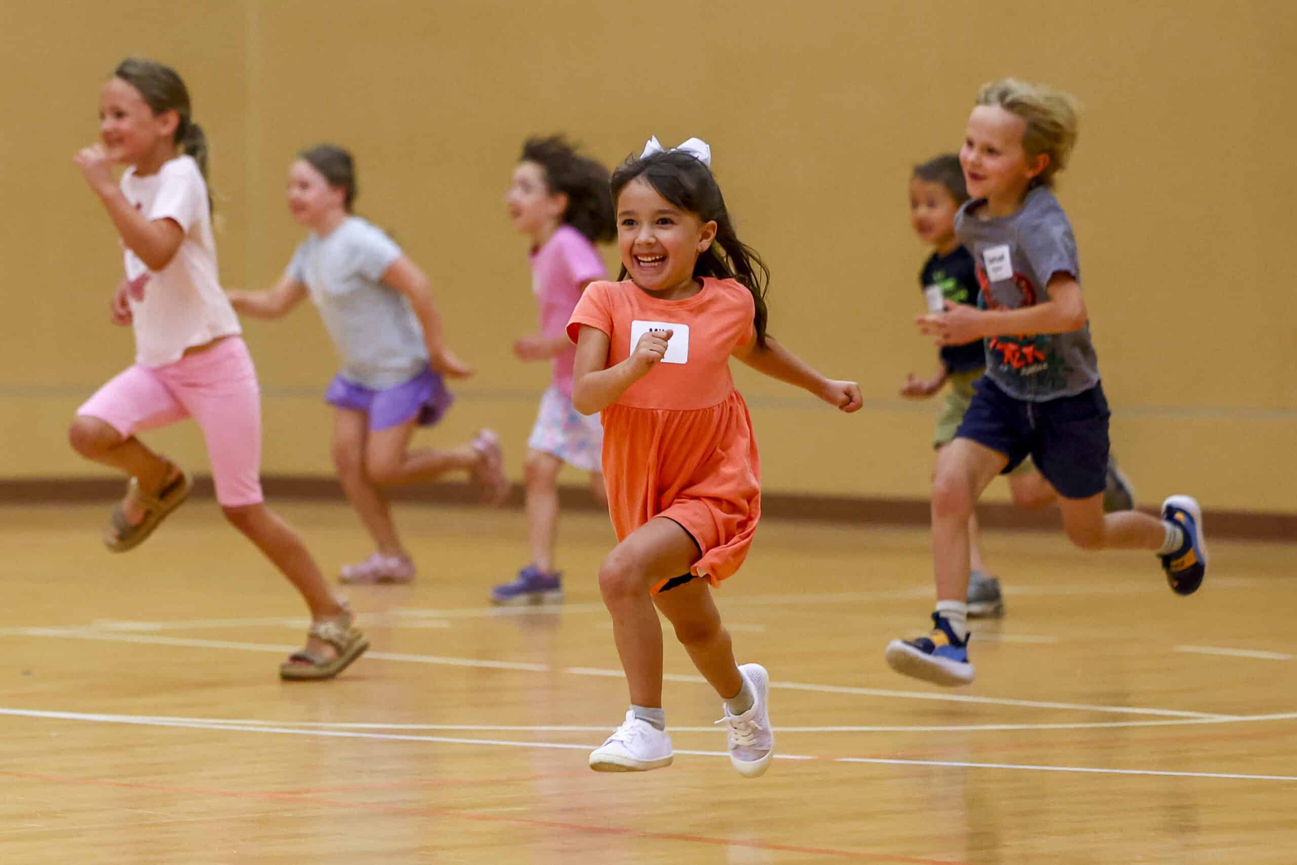 Children indoors holding colorful crafts, standing on a carpeted floor. They appear happy and engaged, with scattered cups on the ground.